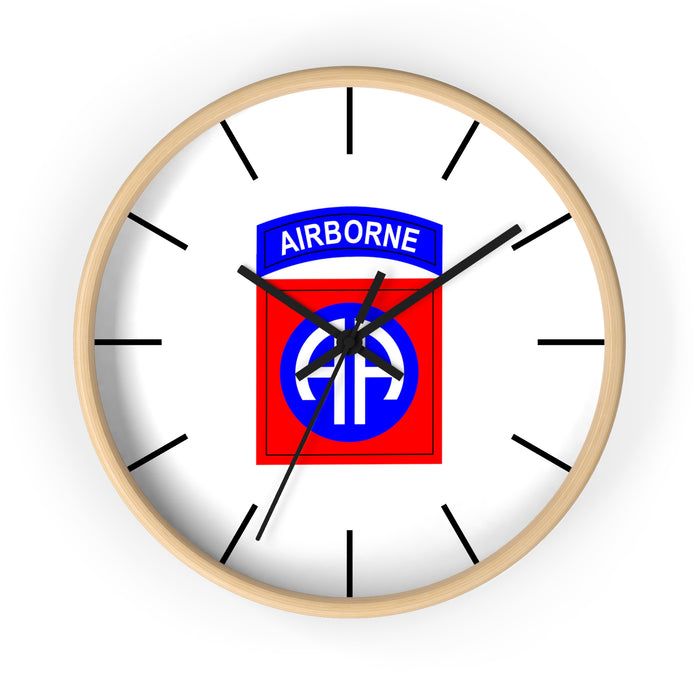 The 82nd Airborne Wall Clock is a round wall clock with a light wood frame and features a white face with black hour and minute marks. At the center of the clock is the red, blue, and white "AIRBORNE" emblem with a winged parachute logo. The time shows approximately 10:08.