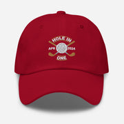 The Golf Hat in cranberry - Hole in One with Personalized Month and Year features a golf design  including two crossed golf clubs with a golf ball at the center and the text "HOLE IN ONE" above. Customized month and year are on the left and right sides.
