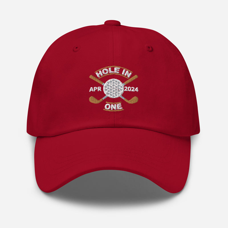 The Golf Hat in cranberry - Hole in One with Personalized Month and Year features a golf design  including two crossed golf clubs with a golf ball at the center and the text "HOLE IN ONE" above. Customized month and year are on the left and right sides.