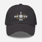 The "I Broke 80" Men's Embroidered Hat is a dark grey, low-profile hat featuring the phrase "I Broke 70" on the front. The word "Broke" is uniquely stylized with a golf ball as the 'O' and crossed golf clubs beneath it.