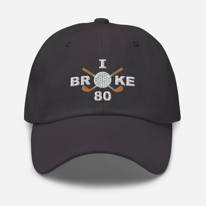 The "I Broke 80" Men's Embroidered Hat is a dark grey, low-profile hat featuring the phrase "I Broke 70" on the front. The word "Broke" is uniquely stylized with a golf ball as the 'O' and crossed golf clubs beneath it.