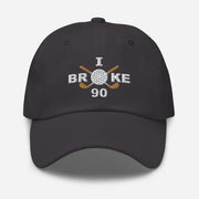 The "I Broke 90" Men's Embroidered Hat is a dark grey, low-profile hat featuring the phrase "I Broke 70" on the front. The word "Broke" is uniquely stylized with a golf ball as the 'O' and crossed golf clubs beneath it.