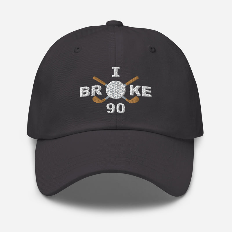 The "I Broke 90" Men's Embroidered Hat is a dark grey, low-profile hat featuring the phrase "I Broke 70" on the front. The word "Broke" is uniquely stylized with a golf ball as the 'O' and crossed golf clubs beneath it.