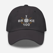 The "I Broke 100" Men's Embroidered Hat is a dark grey, low-profile hat featuring the phrase "I Broke 70" on the front. The word "Broke" is uniquely stylized with a golf ball as the 'O' and crossed golf clubs beneath it.