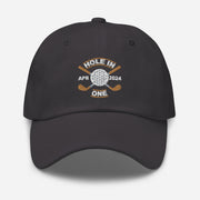 The Golf Hat in dark grey - Hole in One with Personalized Month and Year features a golf design  including two crossed golf clubs with a golf ball at the center and the text "HOLE IN ONE" above. Customized month and year are on the left and right sides.