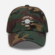 The Golf Hat in green camouflage - Hole in One with Personalized Month and Year features a golf design  including two crossed golf clubs with a golf ball at the center and the text "HOLE IN ONE" above. Customized month and year are on the left and right sides.