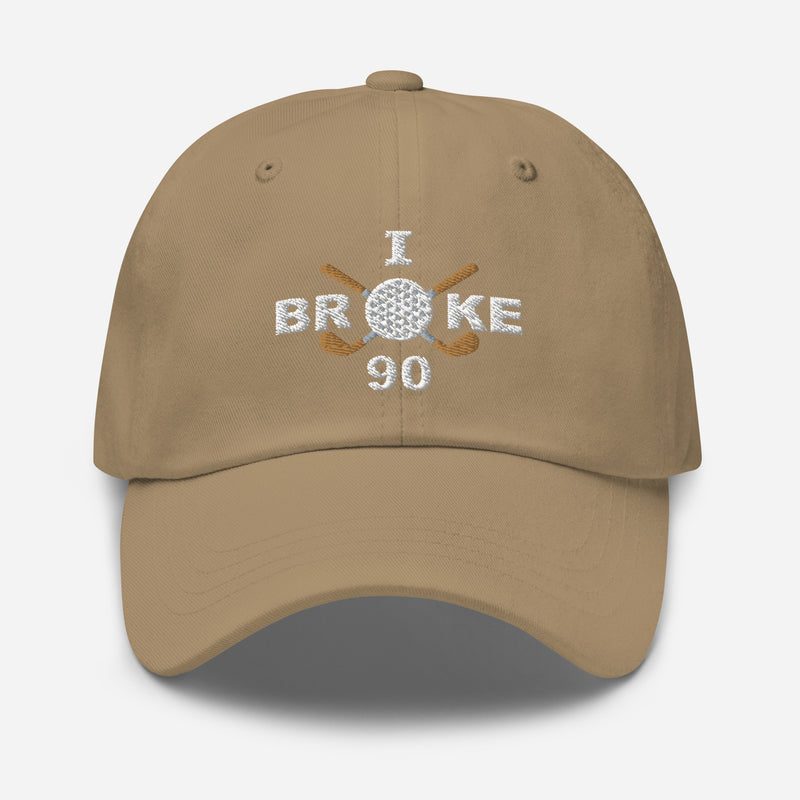 The "I Broke 90" Men's Embroidered Hat is a khaki colored, low-profile hat featuring the phrase "I Broke 70" on the front. The word "Broke" is uniquely stylized with a golf ball as the 'O' and crossed golf clubs beneath it.