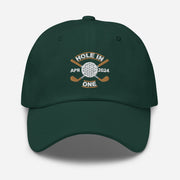 The Golf Hat in spruce colored - Hole in One with Personalized Month and Year features a golf design  including two crossed golf clubs with a golf ball at the center and the text "HOLE IN ONE" above. Customized month and year are on the left and right sides.