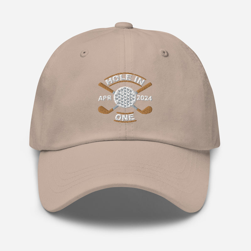 The Golf Hat in stone colored - Hole in One with Personalized Month and Year features a golf design  including two crossed golf clubs with a golf ball at the center and the text "HOLE IN ONE" above. Customized month and year are on the left and right sides.