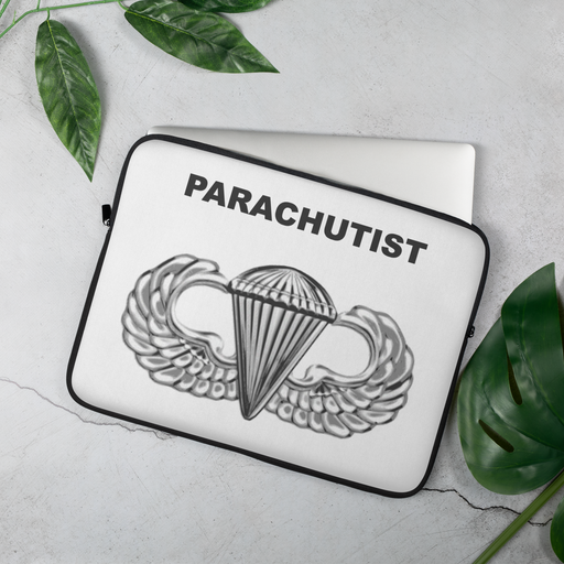 A lightweight Laptop Case - Basic Parachutist with a design featuring a parachutist badge, displaying a parachute flanked by wings and the word "PARACHUTIST" written above it. Its faux fur interior ensures your device stays protected. The case rests on a gray surface surrounded by green leaves.