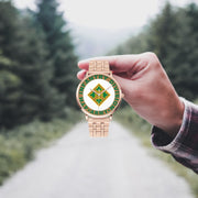 A hand in a plaid shirt holds the 4th Infantry Division Goldtone Watch against the background of a blurred forest scene, where a path runs through the center.