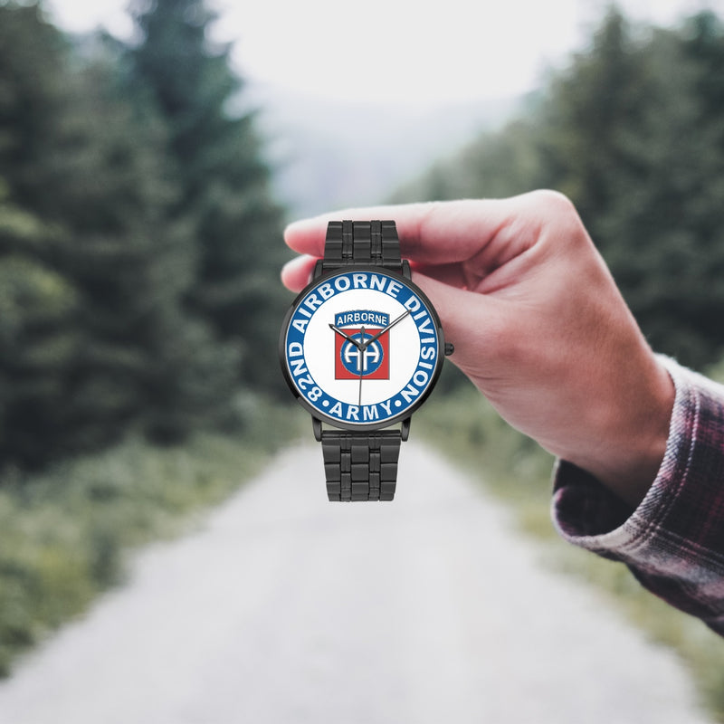 A hand in a plaid shirt holds the 82nd Airborne Division Watch against the background of a blurred forest scene, where a path runs through the center.