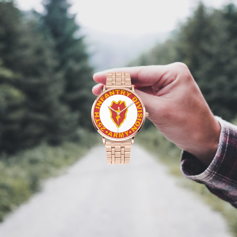 A hand in a plaid shirt holds the 25th Infantry Division Goldtone Watch against the background of a blurred forest scene, where a path runs through the center.