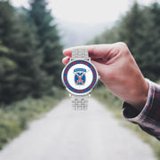 A hand in a plaid shirt holds the 10th Mountain Division Silvertone Watch against the background of a blurred forest scene, where a path runs through the center.