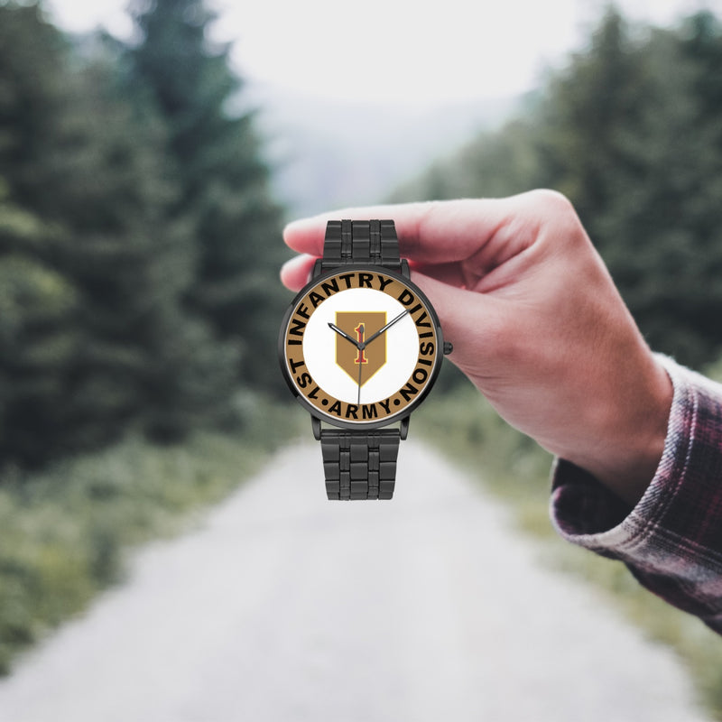 A hand in a plaid shirt holds the 1st Infantry Division Watch against the background of a blurred forest scene, where a path runs through the center.