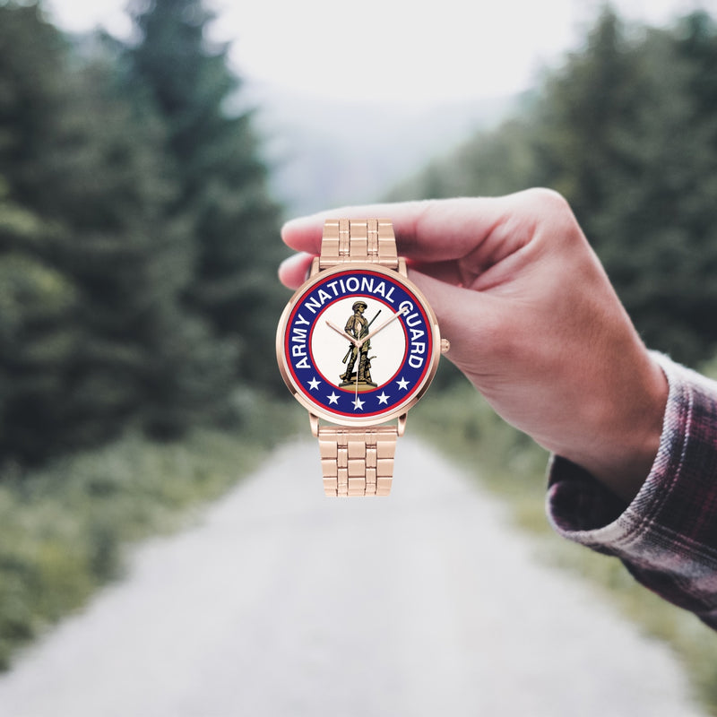 A hand in a plaid shirt holds the US Army National Guard Goldtone Watch against the background of a blurred forest scene, where a path runs through the center.