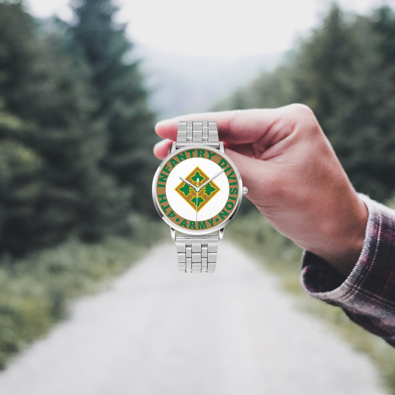 A hand in a plaid shirt holds the 4th Infantry Division Silvertone Watch against the background of a blurred forest scene, where a path runs through the center.