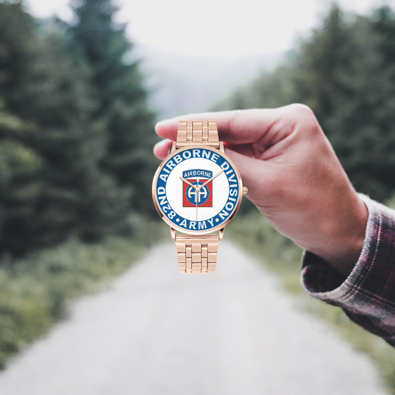 A hand in a plaid shirt holds the 82nd Airborne Division Goldtone Watch against the background of a blurred forest scene, where a path runs through the center.