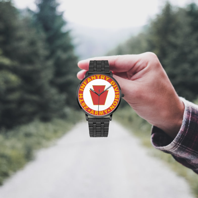 A hand in a plaid shirt holds the 28th Infantry Division Watch against the background of a blurred forest scene, where a path runs through the center.