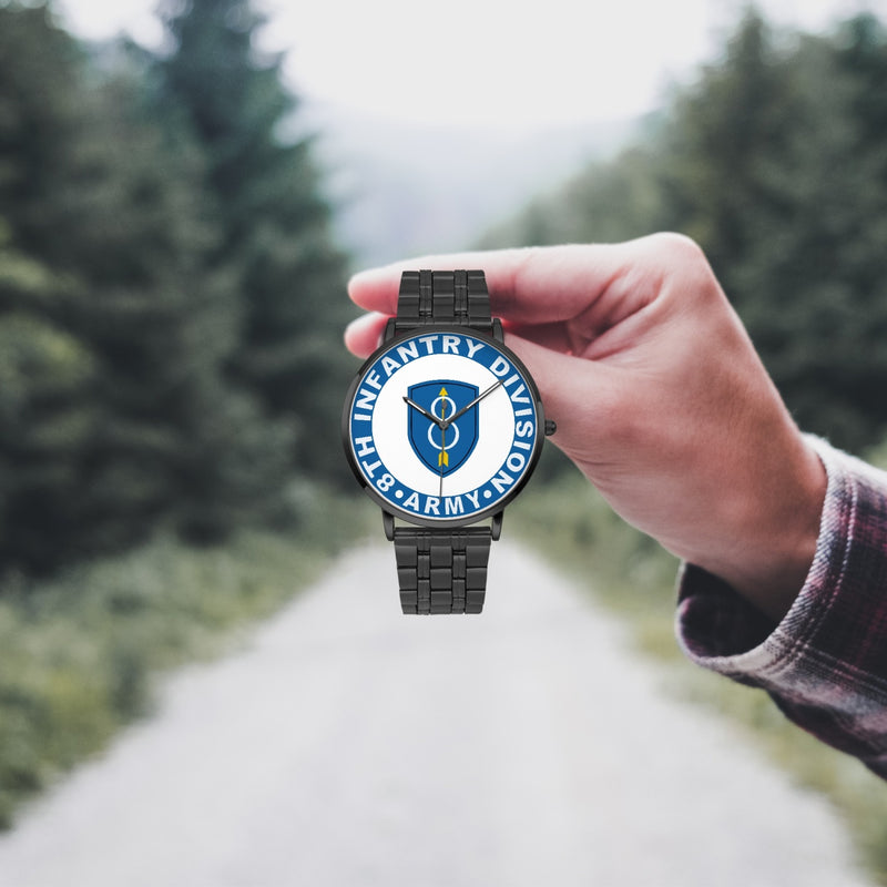 A hand in a plaid shirt holds the 8th Infantry Division Watch against the background of a blurred forest scene, where a path runs through the center.