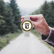 A hand in a plaid shirt holds the 101st Airborne Division Silvertone Watch against the background of a blurred forest scene, where a path runs through the center.
