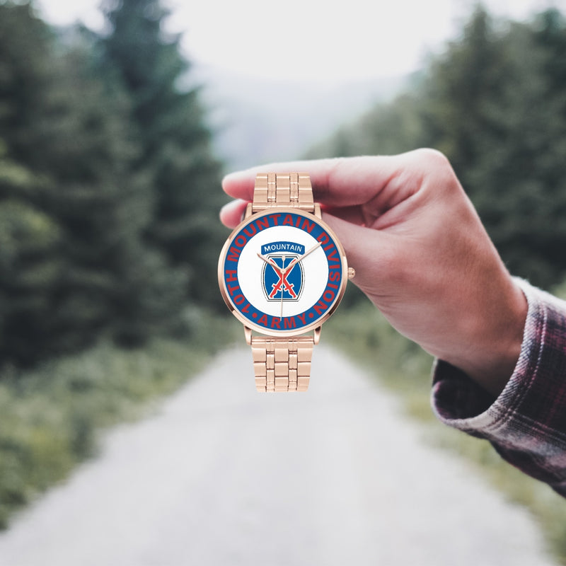 A hand in a plaid shirt holds the 10th Mountain Division Goldtone Watch against the background of a blurred forest scene, where a path runs through the center.