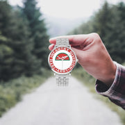 A hand in a plaid shirt holds the 78th Infantry Division Silvertone Watch against the background of a blurred forest scene, where a path runs through the center.
