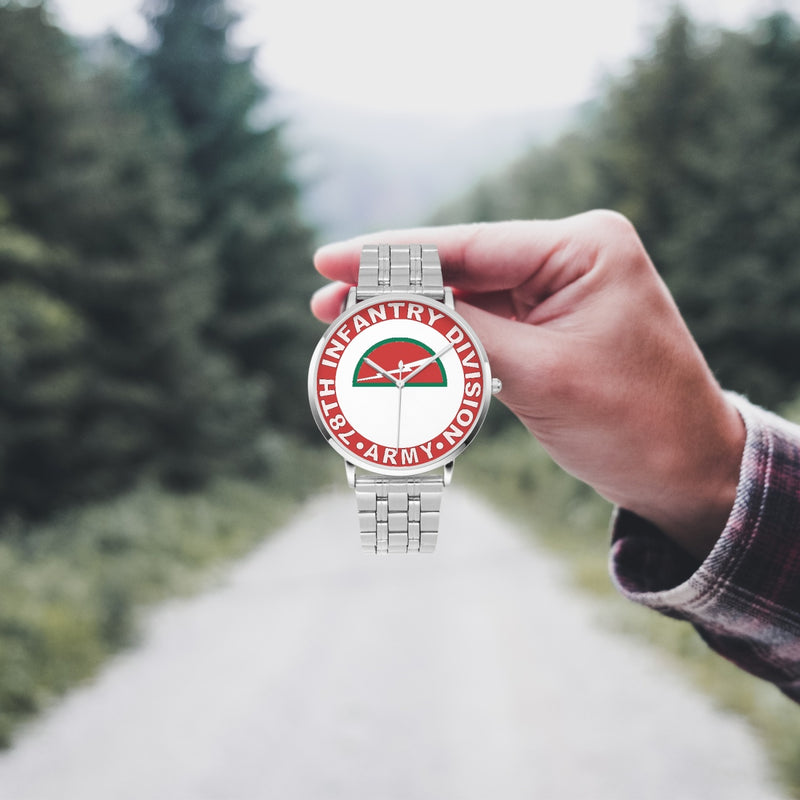 A hand in a plaid shirt holds the 78th Infantry Division Silvertone Watch against the background of a blurred forest scene, where a path runs through the center.