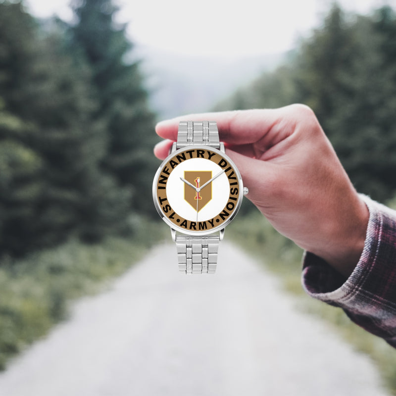 A hand in a plaid shirt holds the 1st Infantry Division Silvertone Watch against the background of a blurred forest scene, where a path runs through the center.
