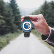 A hand in a plaid shirt holds the 29th Infantry Division Watch against the background of a blurred forest scene, where a path runs through the center.