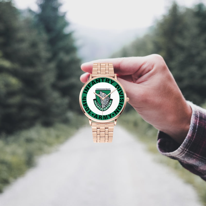 A hand in a plaid shirt holds the 104th Infantry Division Goldtone Watch against the background of a blurred forest scene, where a path runs through the center.