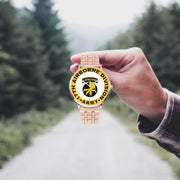 A hand in a plaid shirt holds the 17th Airborne Division Goldtone Watch against the background of a blurred forest scene, where a path runs through the center.