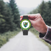 A hand in a plaid shirt holds the 75th Ranger Regiment Watch against the background of a blurred forest scene, where a path runs through the center.