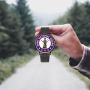 A hand in a plaid shirt holds the US Army National Guard Watch against the background of a blurred forest scene, where a path runs through the center.