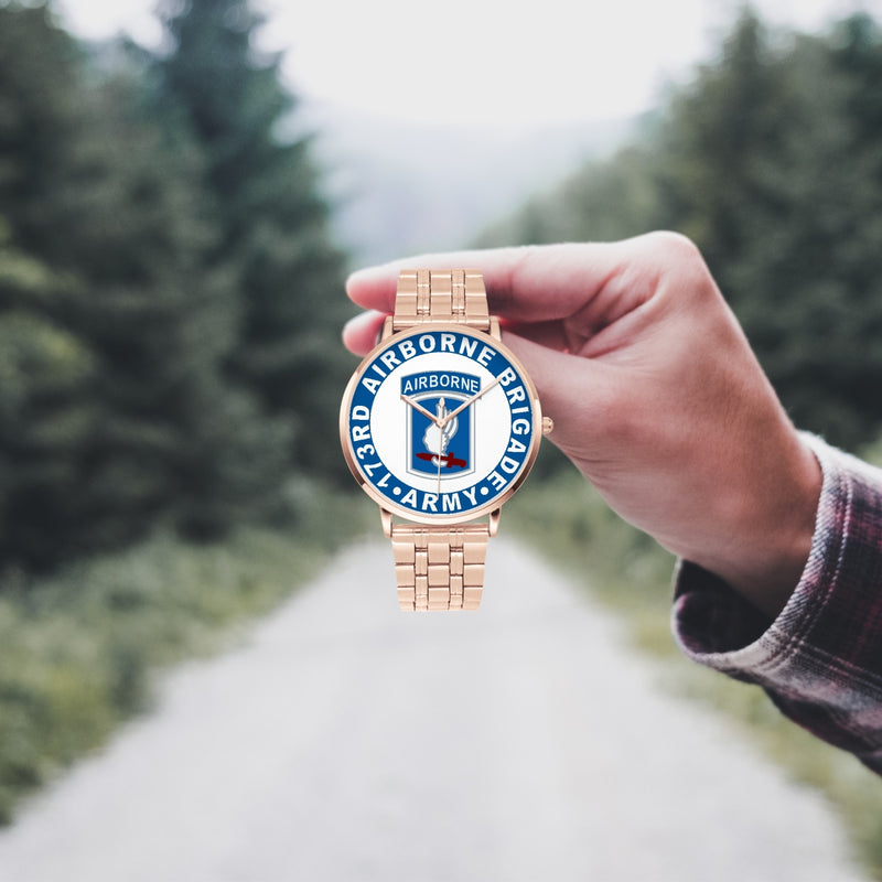 A hand in a plaid shirt holds the 104th Infantry Brigade Goldtone Watch against the background of a blurred forest scene, where a path runs through the center.