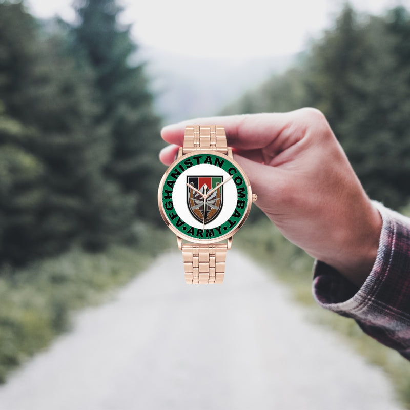 A hand in a plaid shirt holds the US Forces Afghanistan Goldtone Watch against the background of a blurred forest scene, where a path runs through the center.