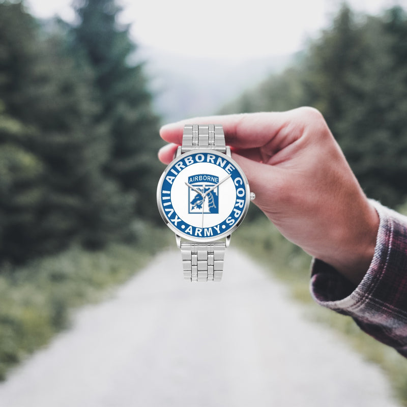 A hand in a plaid shirt holds the XVIII Airborne Corps Silvertone Watch against the background of a blurred forest scene, where a path runs through the center.
