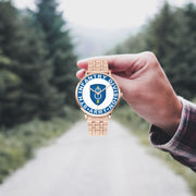 A hand in a plaid shirt holds the 8th Infantry Division Goldtone Watch against the background of a blurred forest scene, where a path runs through the center.