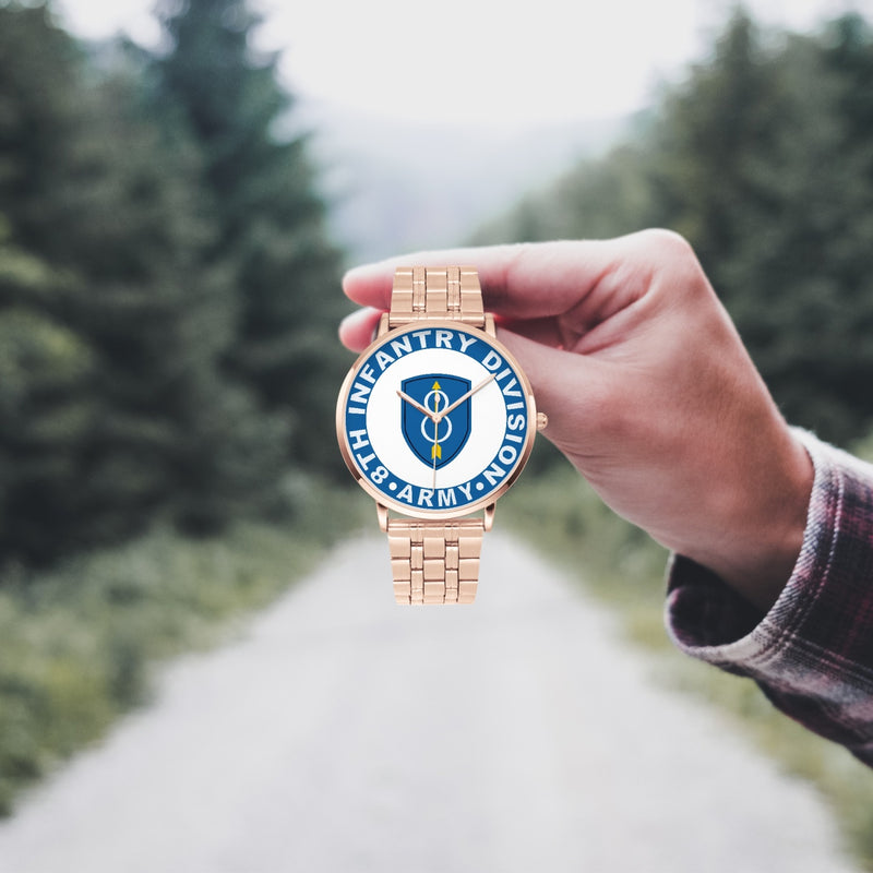 A hand in a plaid shirt holds the 8th Infantry Division Goldtone Watch against the background of a blurred forest scene, where a path runs through the center.