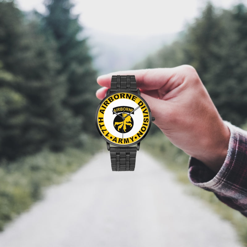 A hand in a plaid shirt holds the 17th Airborne Division Watch against the background of a blurred forest scene, where a path runs through the center.