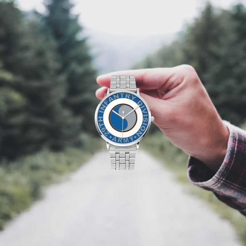 A hand in a plaid shirt holds the 29th Infantry Division Silvertone Watch against the background of a blurred forest scene, where a path runs through the center.