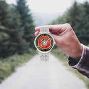 A hand in a plaid shirt holds the United States Marine Corps Silvertone Watch against the background of a blurred forest scene, where a path runs through the center.