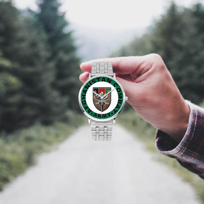 A hand in a plaid shirt holds the US Forces Afghanistan Silvertone Watch against the background of a blurred forest scene, where a path runs through the center.