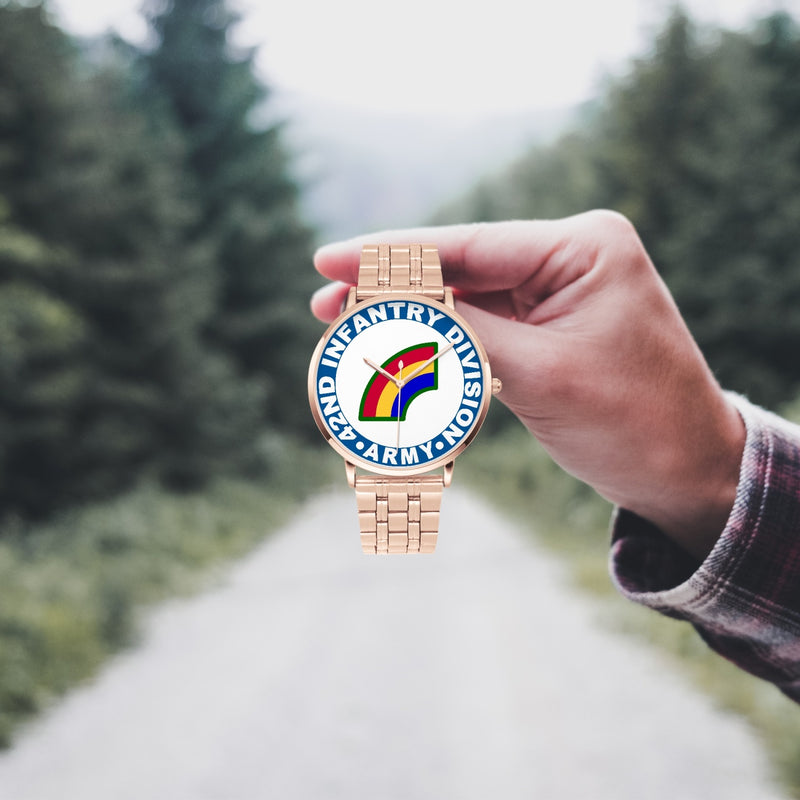 A hand in a plaid shirt holds the 42nd Infantry Division Goldtone Watch against the background of a blurred forest scene, where a path runs through the center.