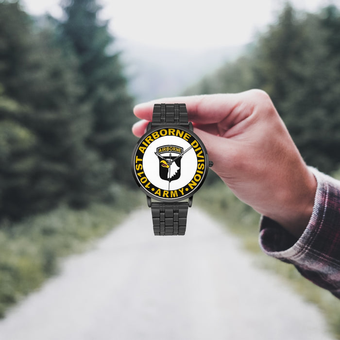 A hand in a plaid shirt holds the 101st Airborne Division Watch against the background of a blurred forest scene, where a path runs through the center.