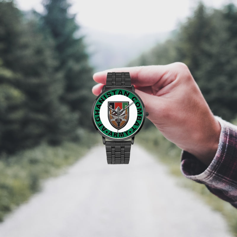 A hand in a plaid shirt holds the US Forces Afghanistan Watch against the background of a blurred forest scene, where a path runs through the center.