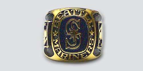 A gold colored paperweight featuring the Seattle Mariners logo set against a black enamel background. The words "Seattle Mariners" are engraved on the bezel.