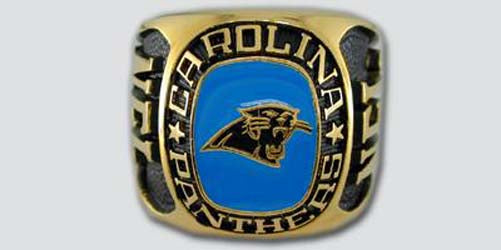 A gold colored paperweight featuring the Carolina Panthers logo set against a blue enamel background. The words "Carolina Panthers" are engraved on the bezel.