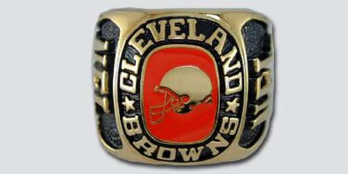 A gold colored paperweight featuring the Cleveland Browns logo set against an orange enamel background. The words "Cleveland Browns" are engraved on the bezel.
