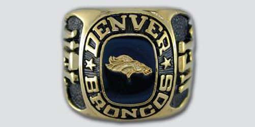 A gold colored paperweight featuring the Denver Broncos logo set against a dark blue enamel background. The words "Denver Broncos" are engraved on the bezel.
